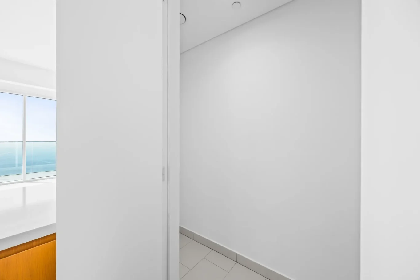Bright white interior: doorway into a small hallway with tiled floor; left reveals a kitchen counter and large window overlooking the sea.