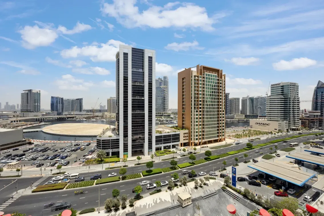 A modern city skyline with tall glass buildings along a waterway and busy roads below under a blue sky.
