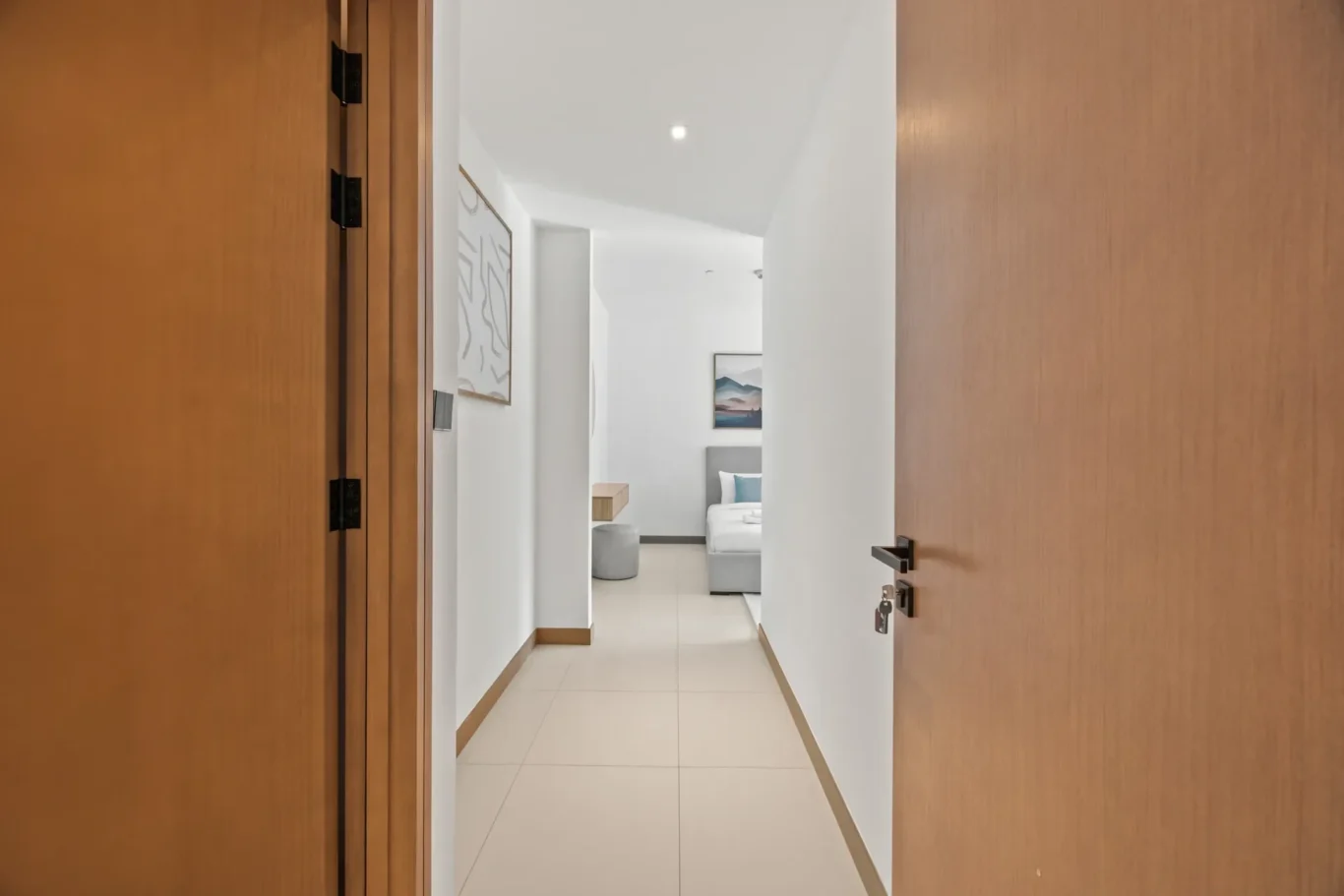 View down a bright hallway into a modern bedroom with a bed and blue pillows at the far end, framed by wooden doorways on both sides