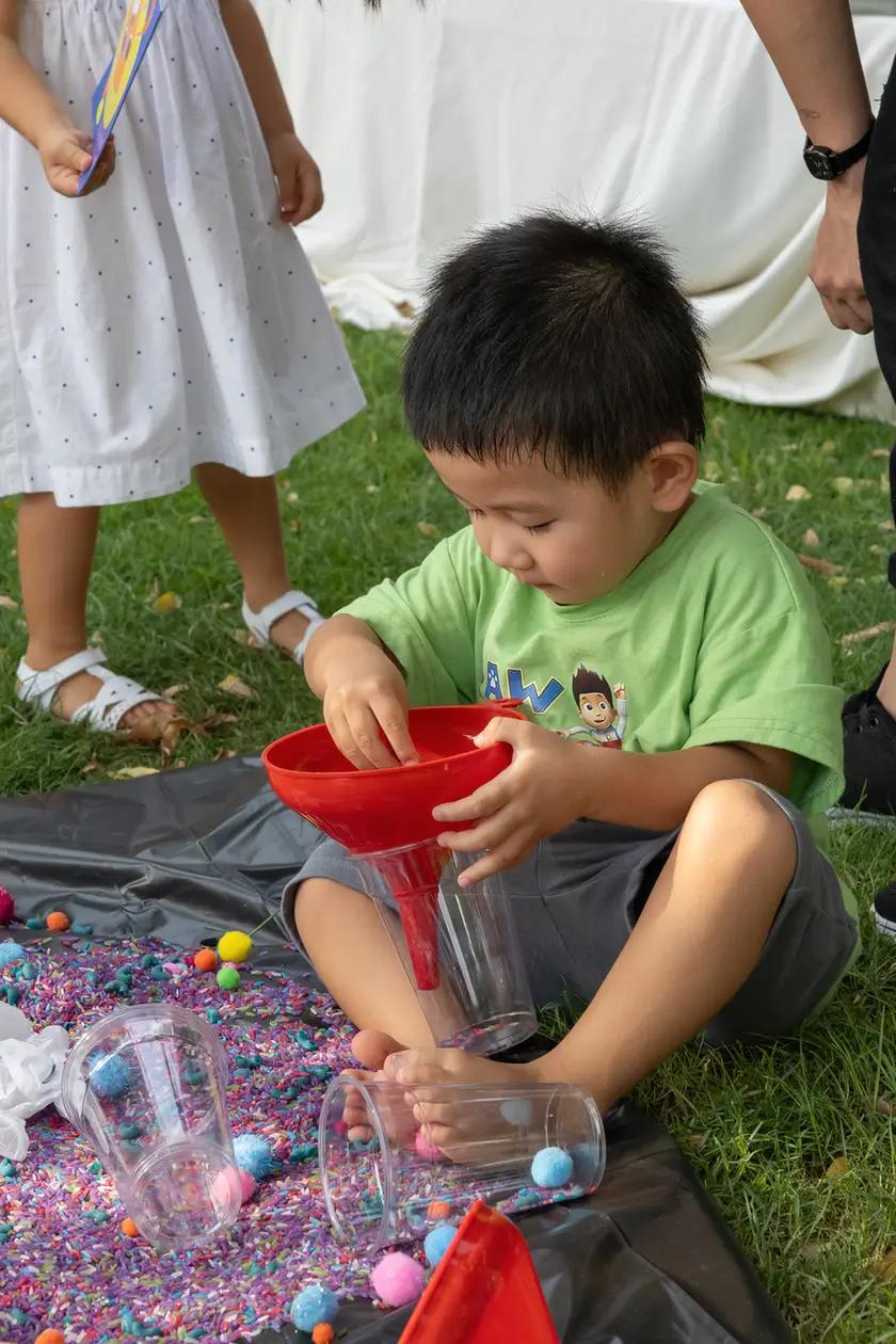 Young boy sitting on grass, pouring beads through a red funnel into clear cups on a colorful bead-filled mat outdoors.