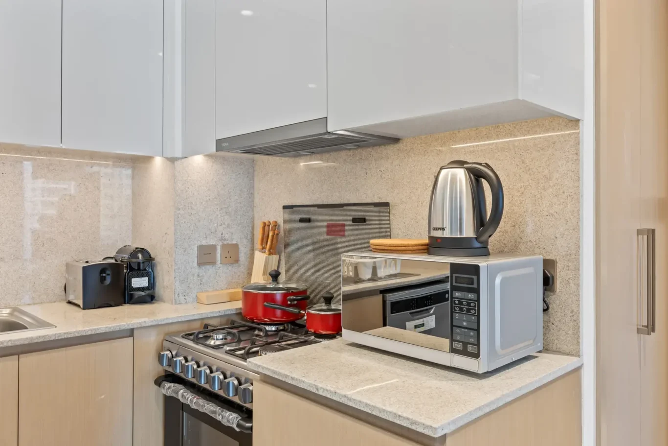 Modern kitchen counter with a stainless microwave, electric kettle, red pots on the stove, and beige granite surfaces.