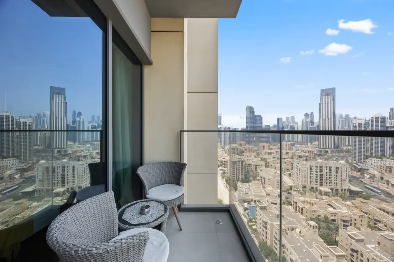 Balcony seating area with wicker chairs and a small round table, glass railing, overlooking a dense city skyline of skyscrapers under a blue sky.