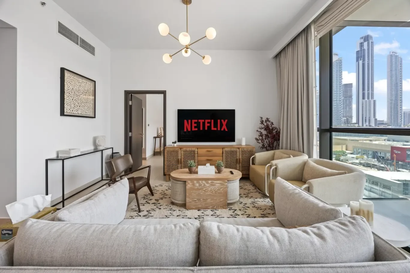 Modern living room with beige sofas, a round coffee table, and a TV showing Netflix, near a large window with a city skyline outside.