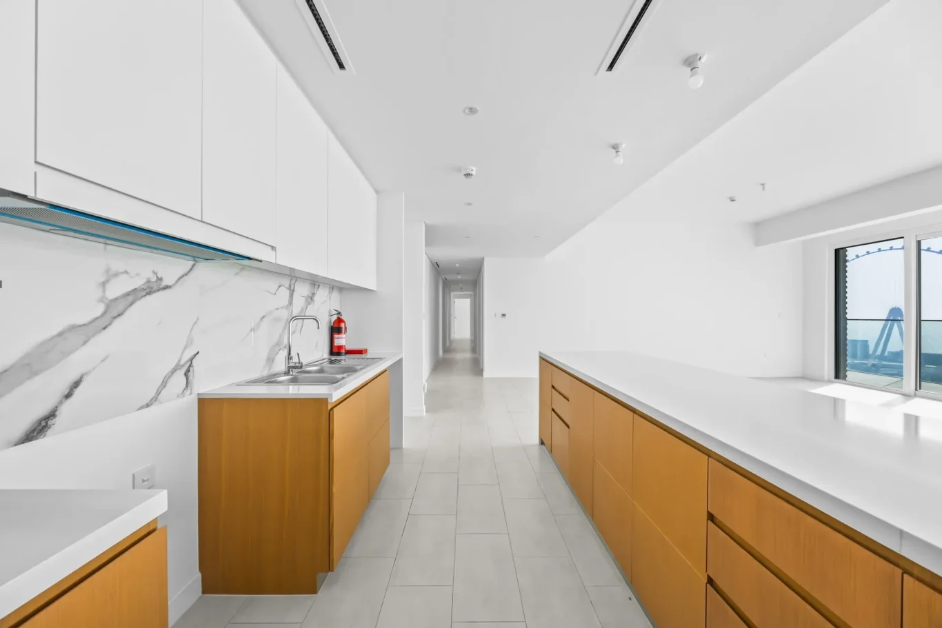 Bright modern kitchen with white upper cabinets, wood-toned lower cabinets, and a marble backsplash, extending into a hallway.