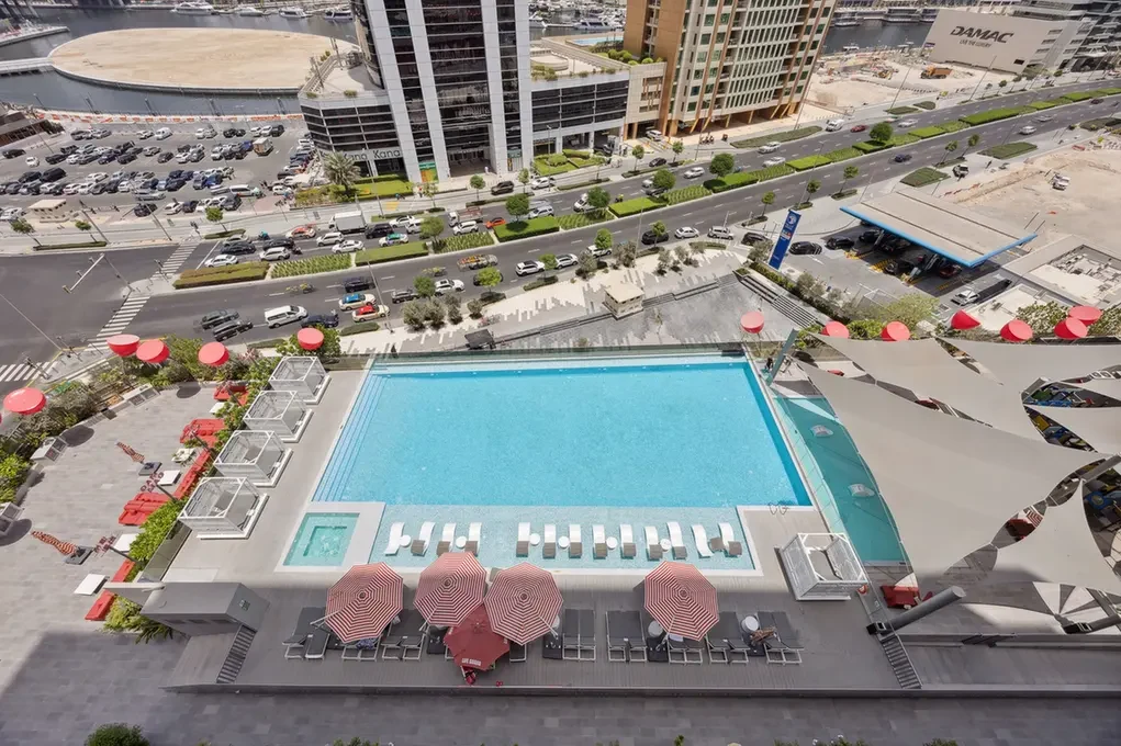A rooftop resort pool area with red umbrellas and lounge chairs, overlooking a city street.