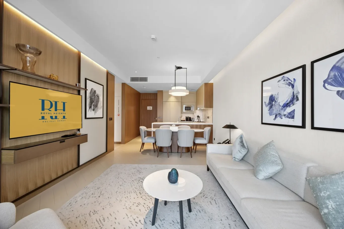 Open-plan living space with a beige sofa, blue cushions, and a white coffee table featuring a teal vase, facing a wall TV showing a yellow Royal Heights logo.