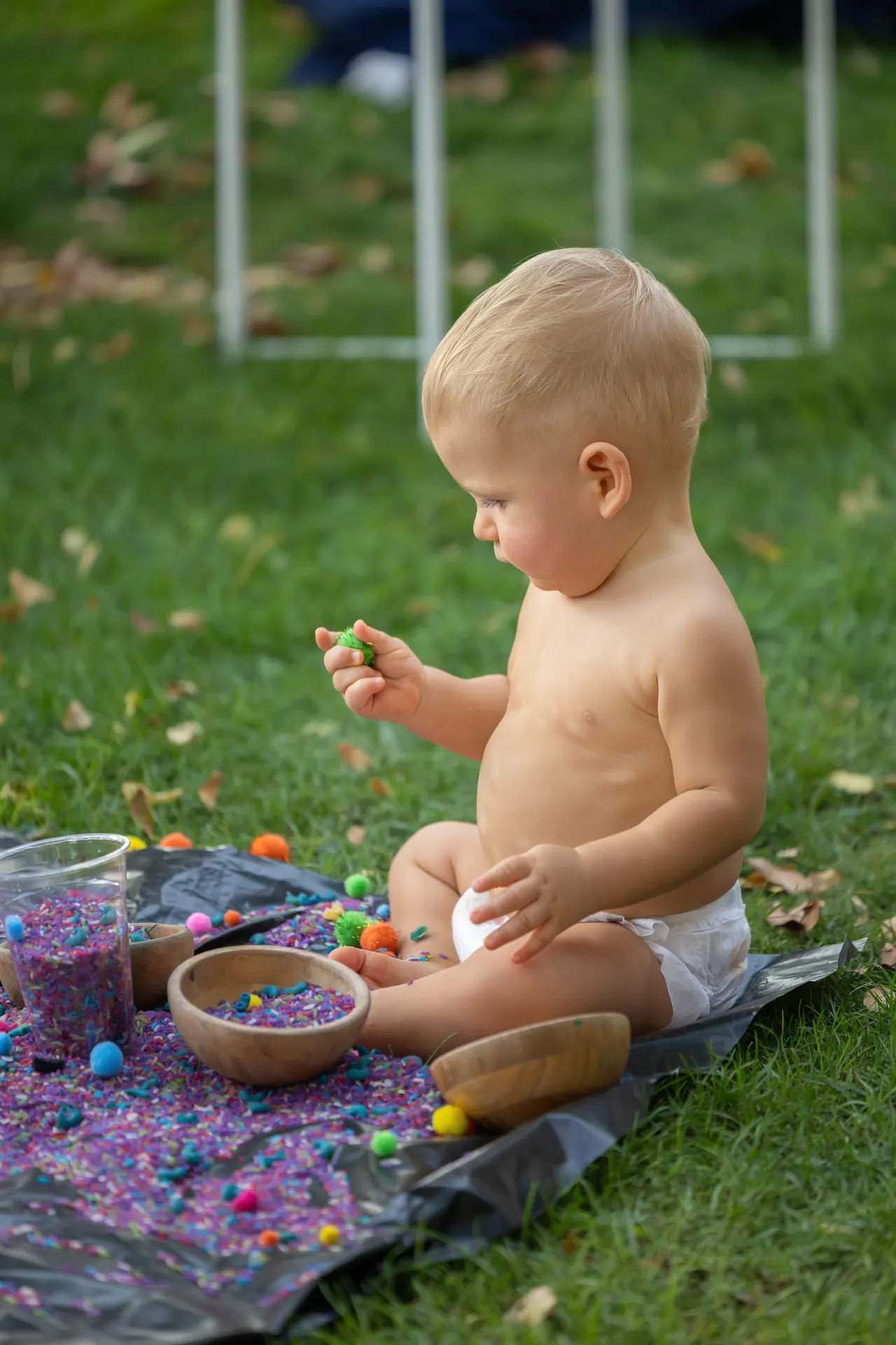 Toddler sits on a blanket in a park, playing with colorful beads and small bowls scattered around.
