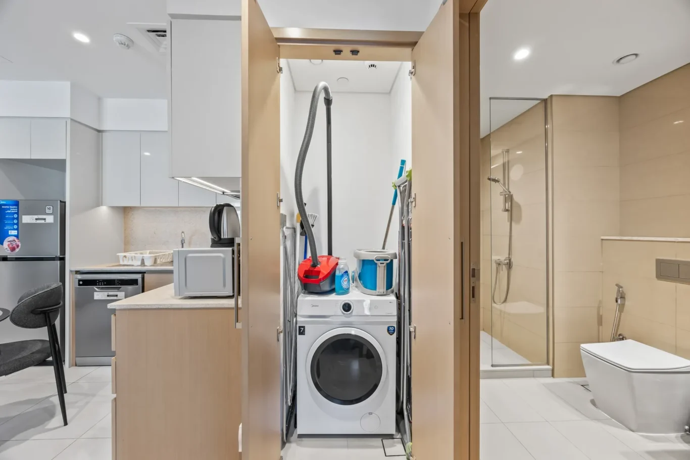 Open laundry closet between the kitchen and bathroom showing a stacked washer and dryer with a red vacuum, mop, and cleaning supplies inside.