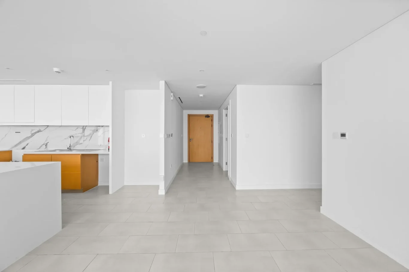 Bright, empty hallway in a modern home with a wooden door at the end and a white kitchen on the left.
