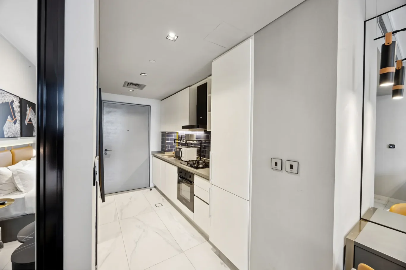 Modern galley kitchen with white cabinets, black range hood, stove and microwave along a gray-white tile floor corridor view down the room.