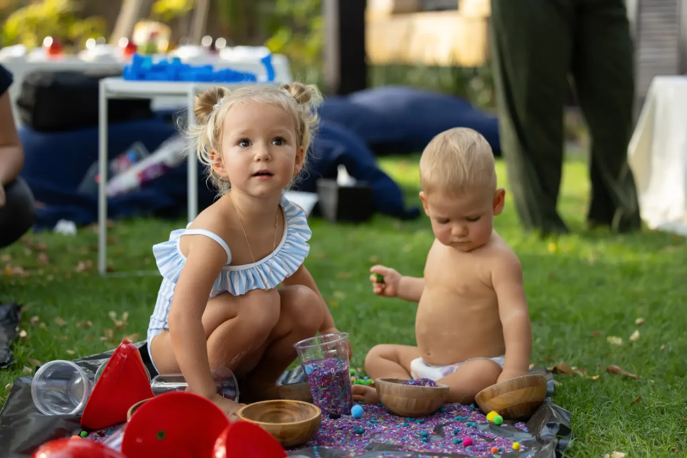 Two young children sit on a blanket in a park, playing with colorful beads and bowls during a sunny outdoor gathering.