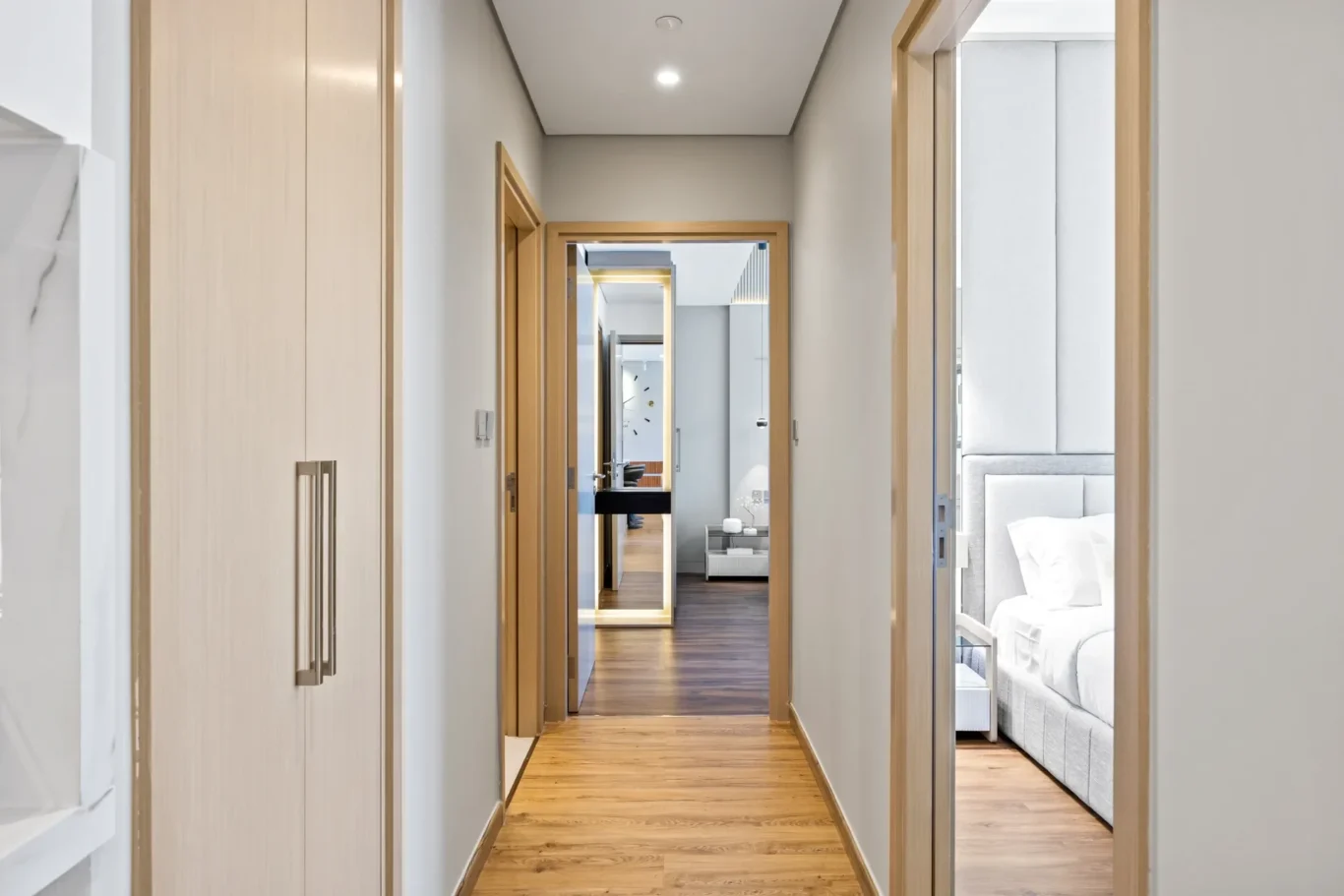 Narrow hallway in a modern home with light wood doors and a hardwood floor, leading to a bedroom at the end.