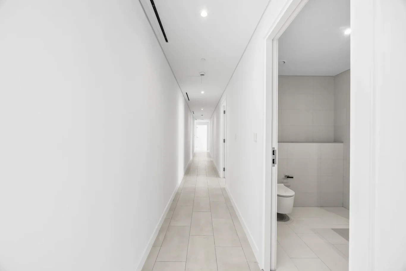 Bright white hallway with tiled floor and multiple doors, bathroom visible on the right with toilet and sink beyond the doorway far end of the corridor.