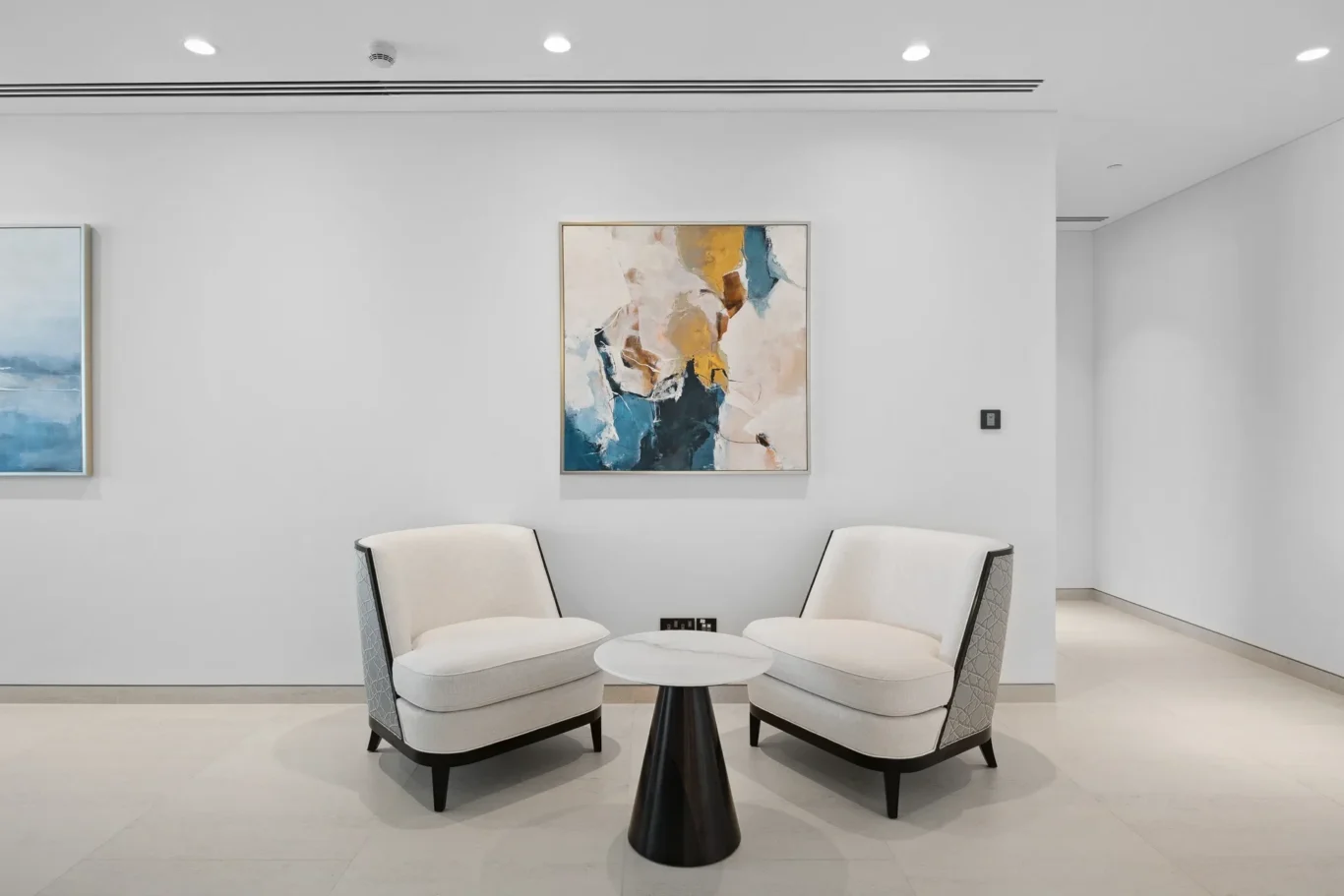 Modern lounge with two cream curved chairs facing a small black pedestal table, abstract painting on the wall between them.
