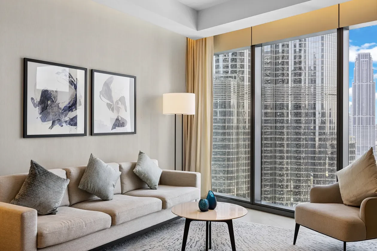 Contemporary living room with a beige sofa and multiple gray cushions, a round coffee table, floor lamp, abstract art, and floor-to-ceiling city windows.