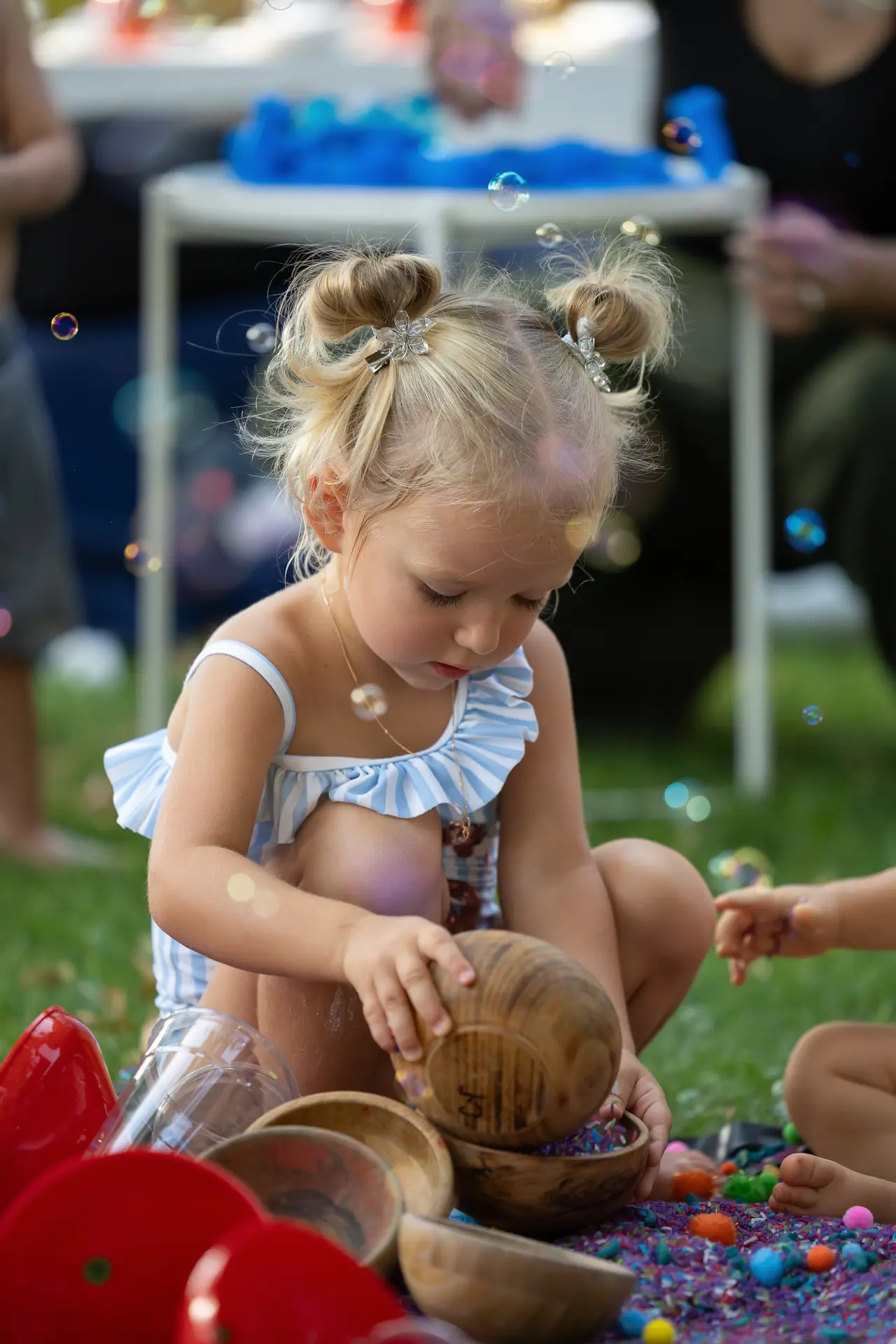 Young girl with blonde hair in buns playing with wooden bowls and colorful beads on the grass outdoors, bubbles floating around.