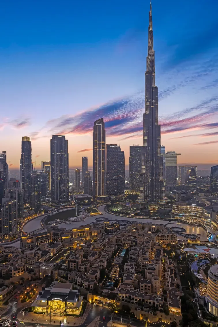 Aerial view of Dubai skyline at dusk with Burj Khalifa towering above modern glass buildings and a lit urban complex below.