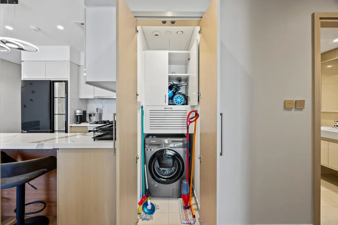 Narrow built-in laundry cabinet in a modern kitchen, doors open to reveal a front-loading washing machine at the bottom with cleaning supplies and mop tools on shelves above.