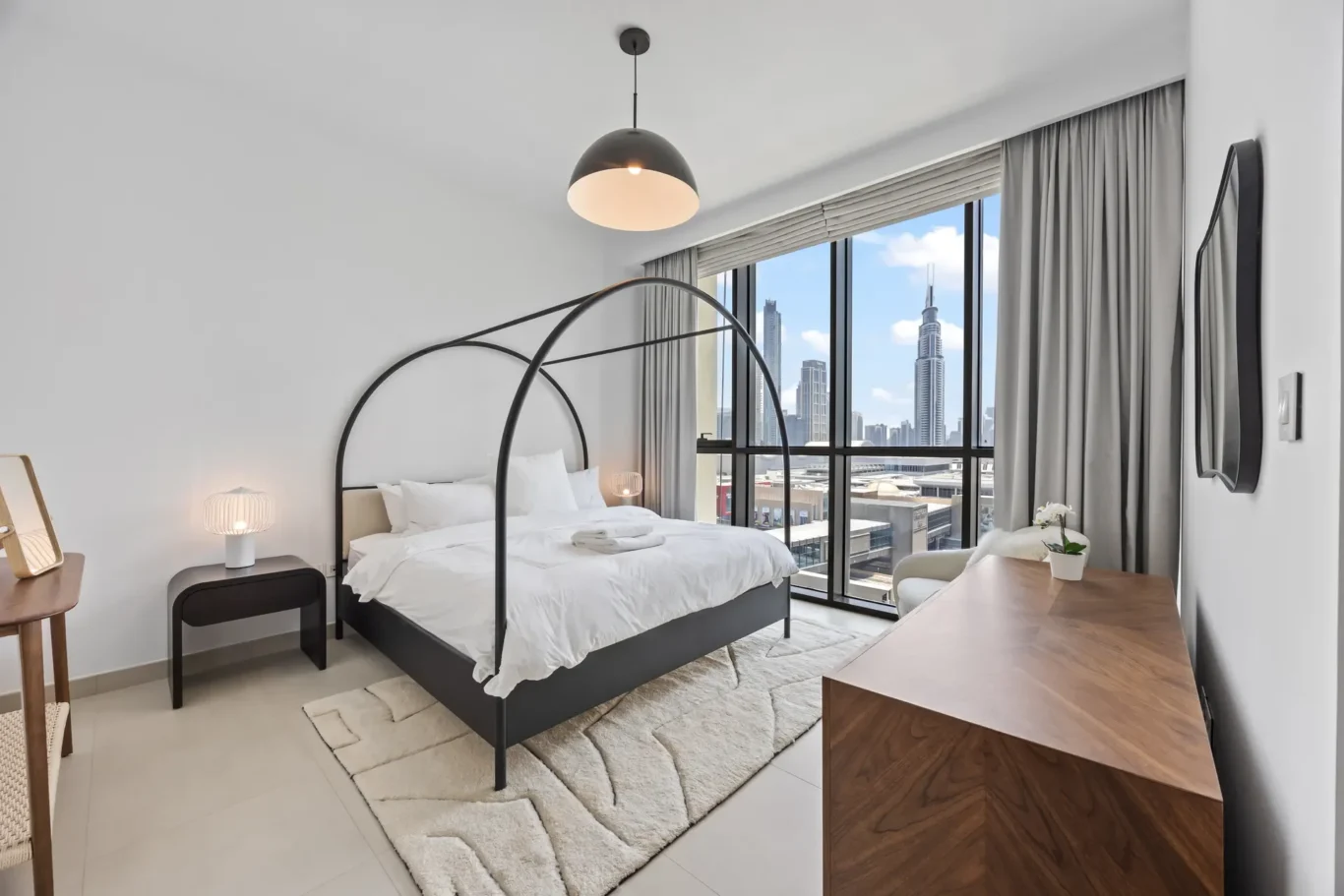 Hotel bedroom with a queen bed, black metal arches, bedside lamps, and a large window with city skyline.