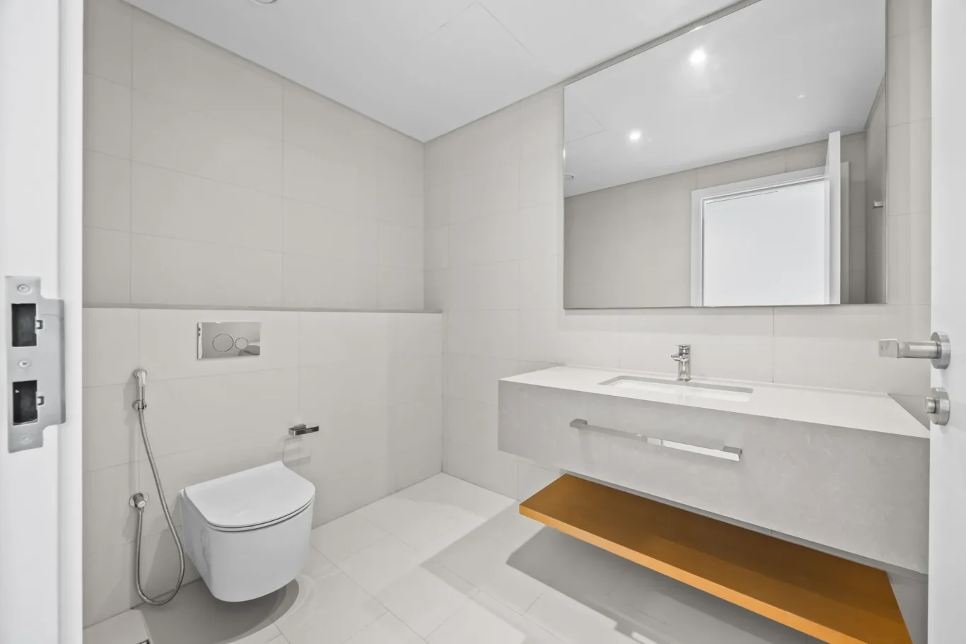 Modern white bathroom with wall-mounted sink, large mirror, and floating wood shelf; toilet and bidet hose visible; open door on left