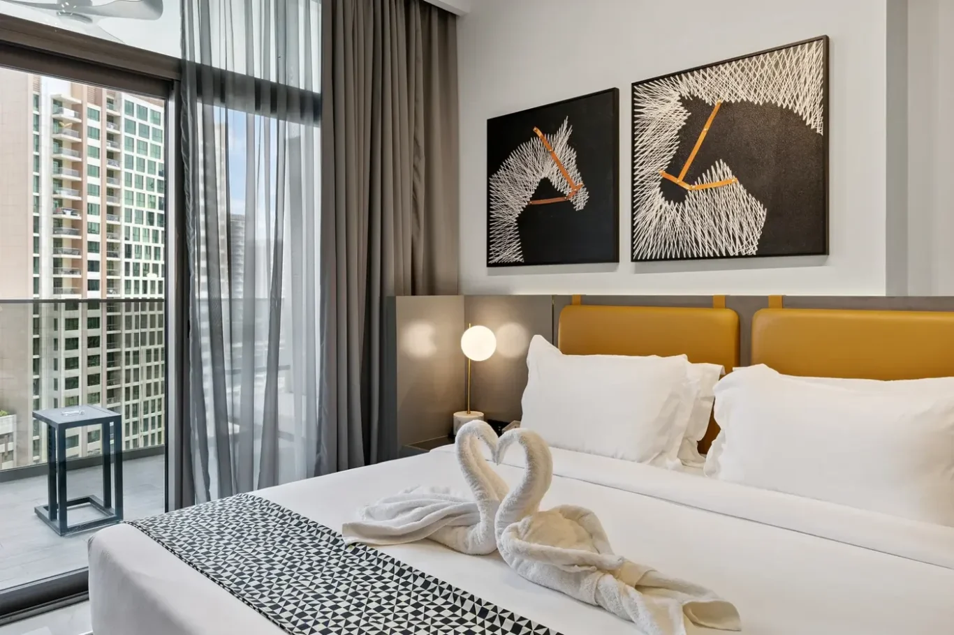 Hotel room with white bedding and a yellow headboard, swan towels on the bed, and abstract art above. Balcony and city view visible outside the glass doors.