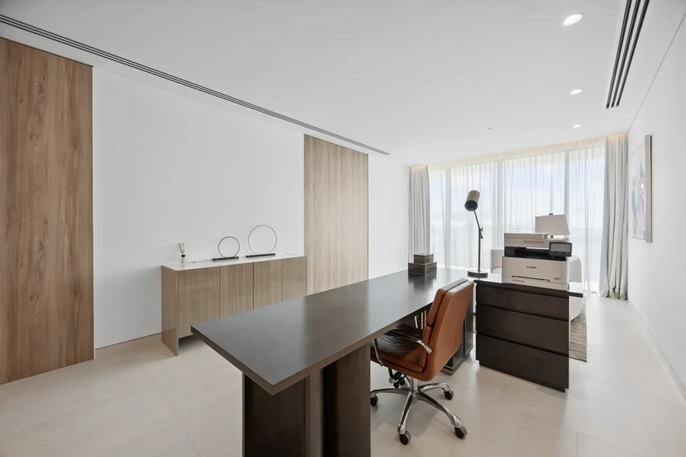 Modern office with a long dark desk, brown ergonomic chair, wood-paneled cabinets, and a Canon printer against a bright window wall.