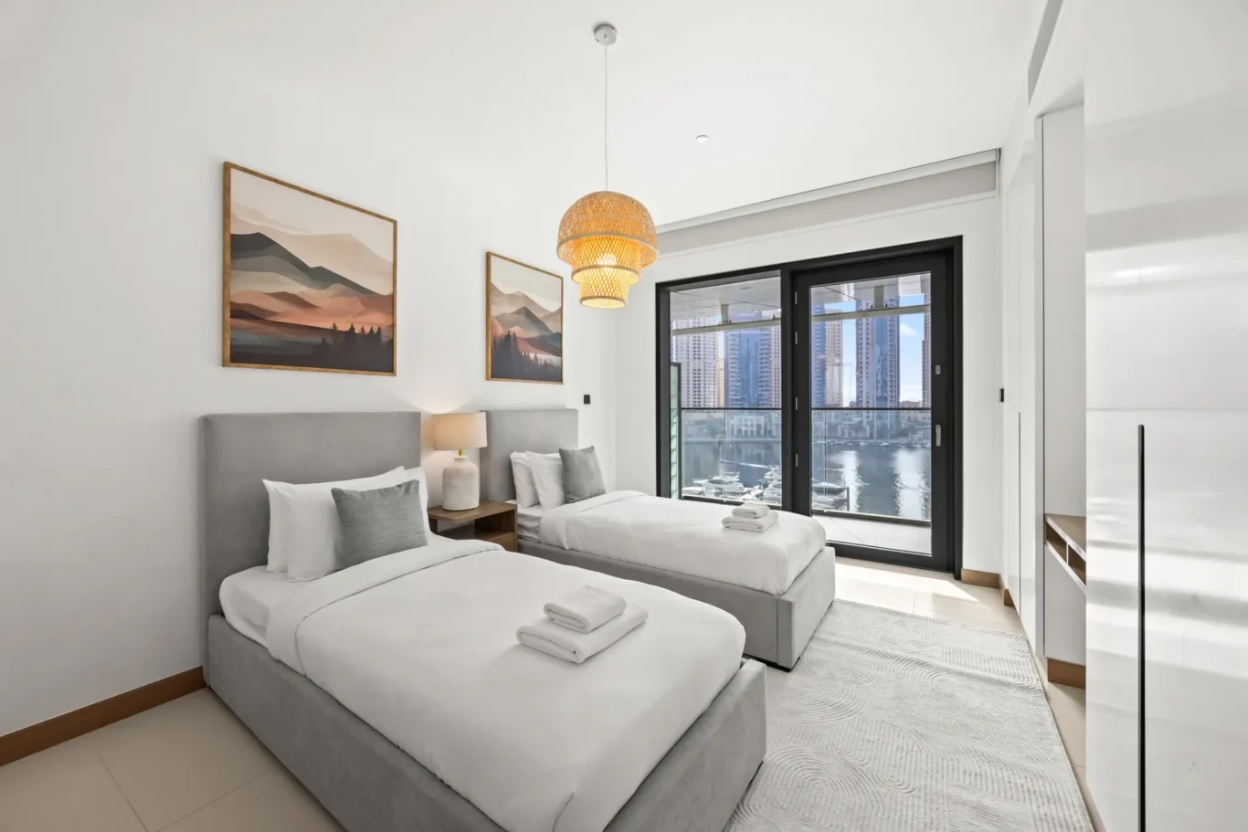 Twin-bedroom in a modern hotel with two gray upholstered beds, white bedding, a nightstand lamp, wall art, and a large window overlooking water and city buildings.