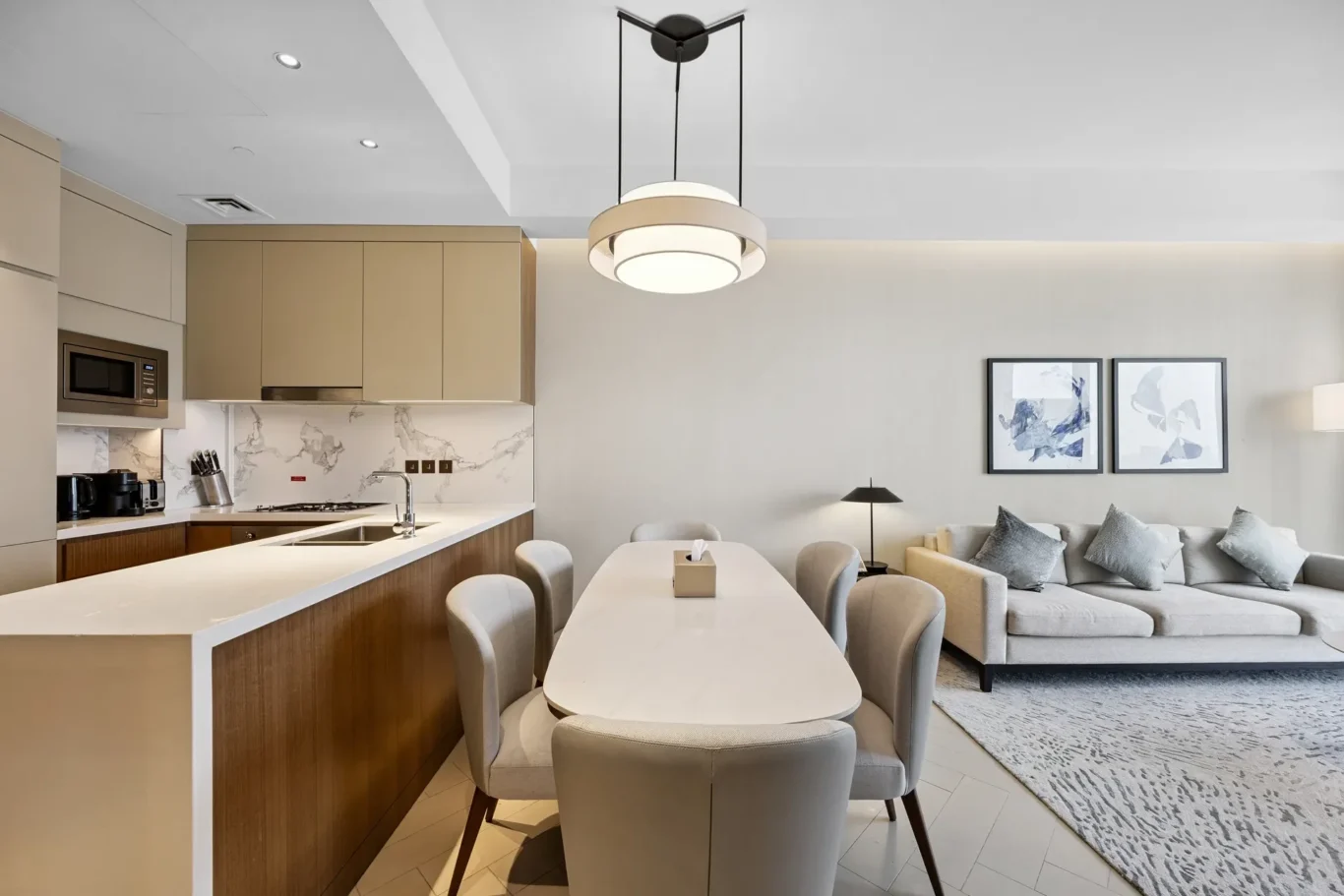 Open-plan kitchen and living area with beige cabinets, marble backsplash, white island, and an oval dining table with gray chairs.