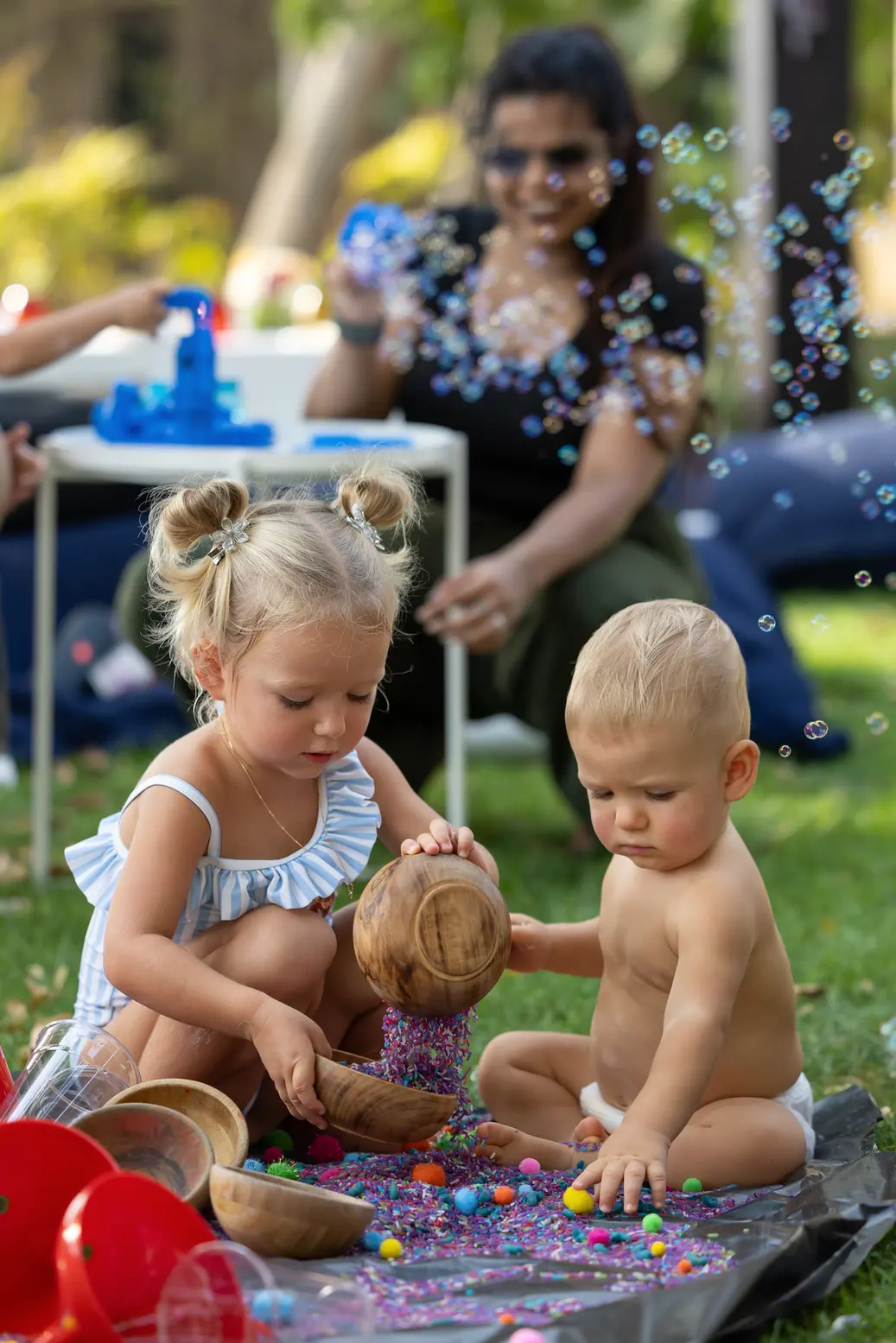 Two young children play with colorful beads and wooden scoops on a tarp in a sunny yard, bubbles floating around as an adult watches nearby.