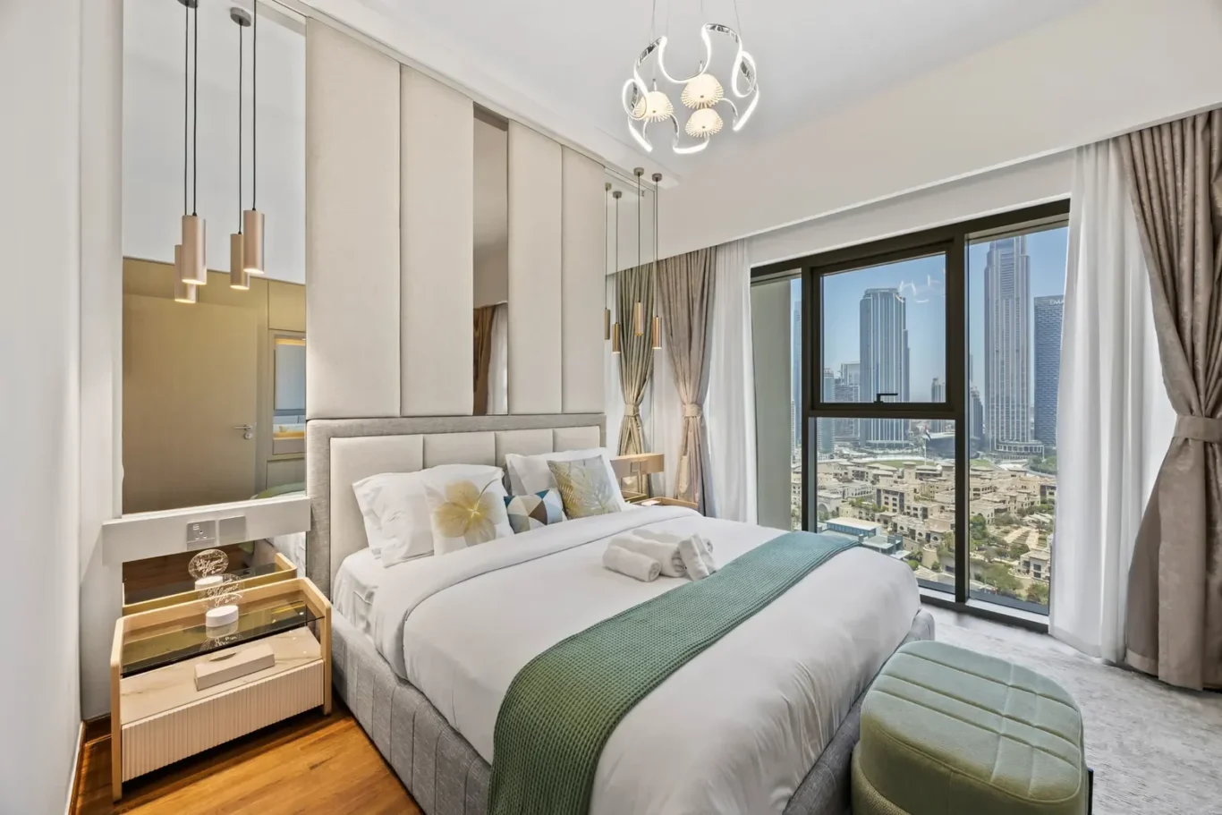 Luxury hotel-style bedroom with a large window showing a city skyline, a plush bed with white linens and teal throw, and modern pendant lights above a nightstand.