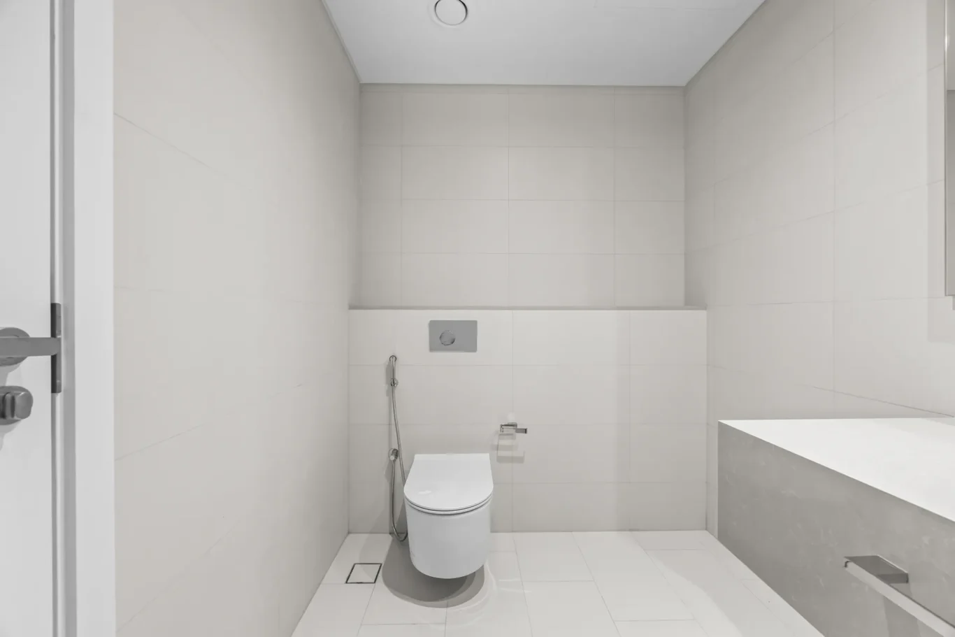 Modern minimalist bathroom with a wall-mounted toilet, handheld bidet spray, and a wall flush plate amid white tiled walls and floor.