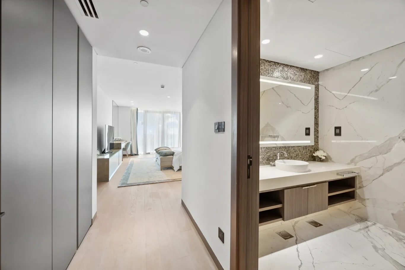Studio suite with en suite marble bathroom and wooden vanity seen through a doorway; light-filled bedroom area in the background.