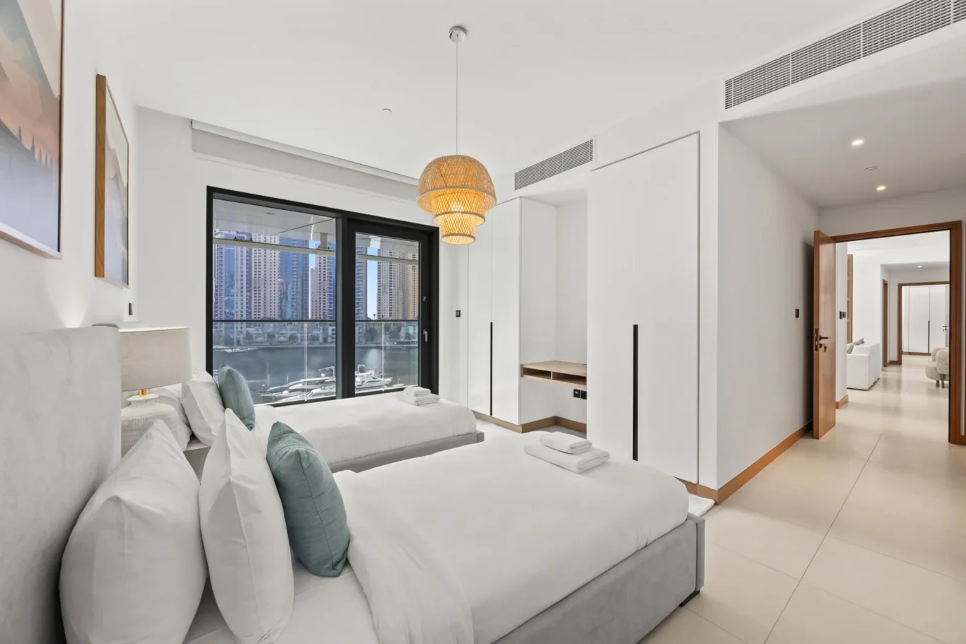 Bright hotel bedroom with two white beds, teal pillows, and a large window overlooking the city skyline and marina; woven pendant light overhead.