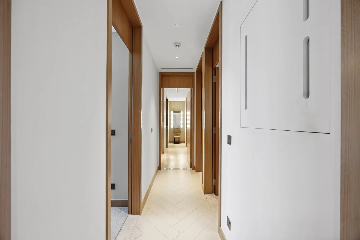 Bright, modern hallway with white walls and wooden door frames, multiple doors along a long corridor, tiled floor, and ceiling lights.
