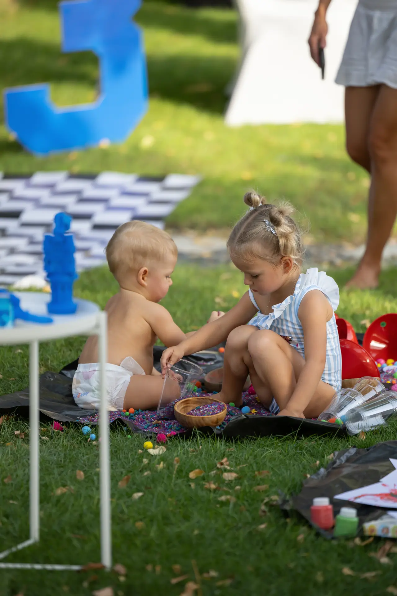 Two toddlers play with colorful beads on a blanket in a park, supervised by adults nearby, with a large blue number 3 in the background.