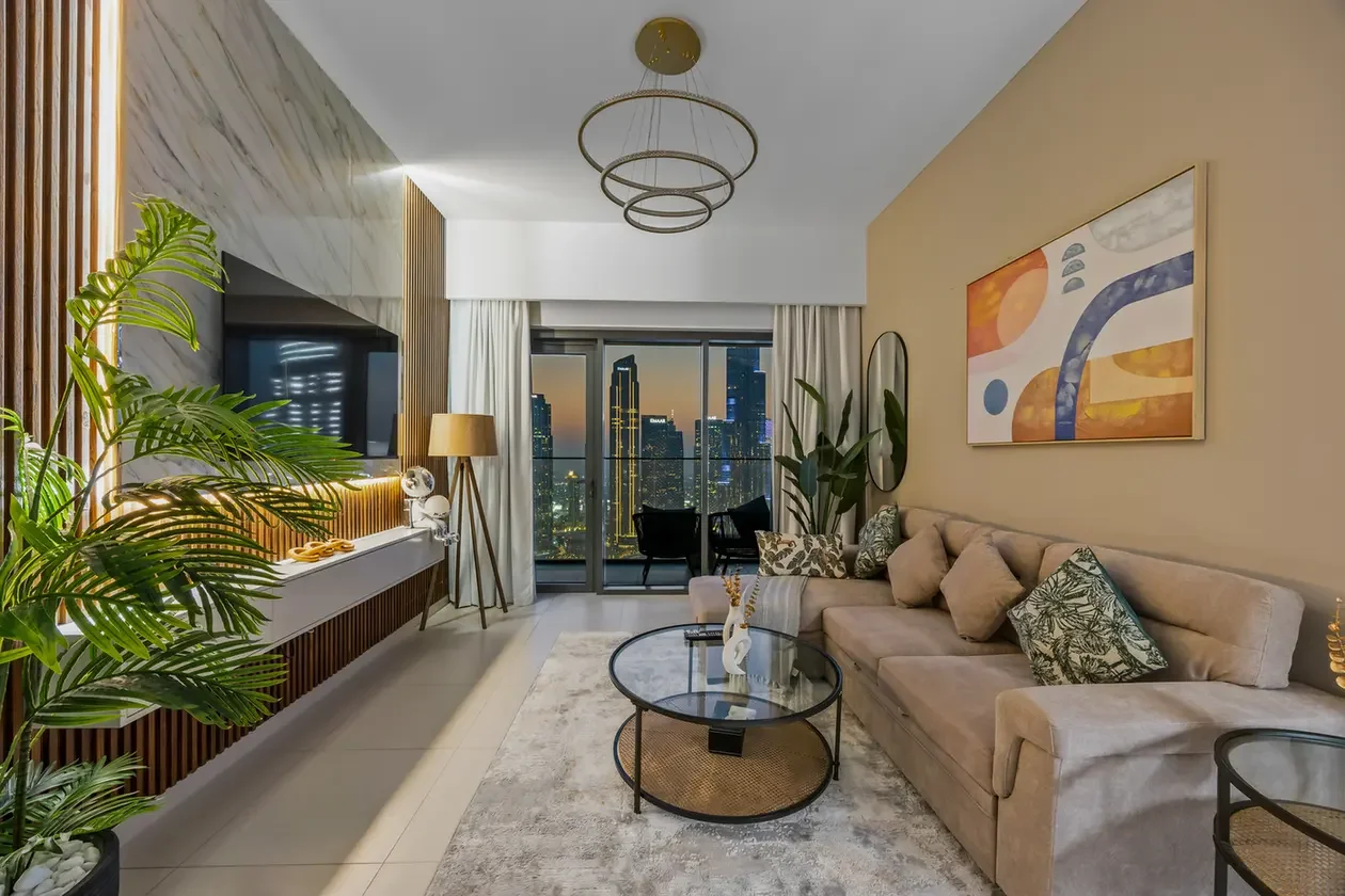 Modern living room with a beige sectional sofa, glass coffee table, and abstract wall art, opening to a city skyline balcony at dusk.