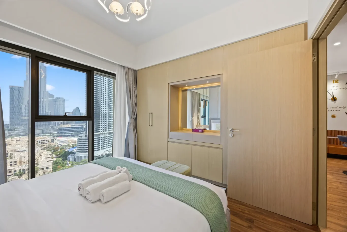 Hotel bedroom with a large window showing city skyline, bed with rolled towels and a green bed runner.