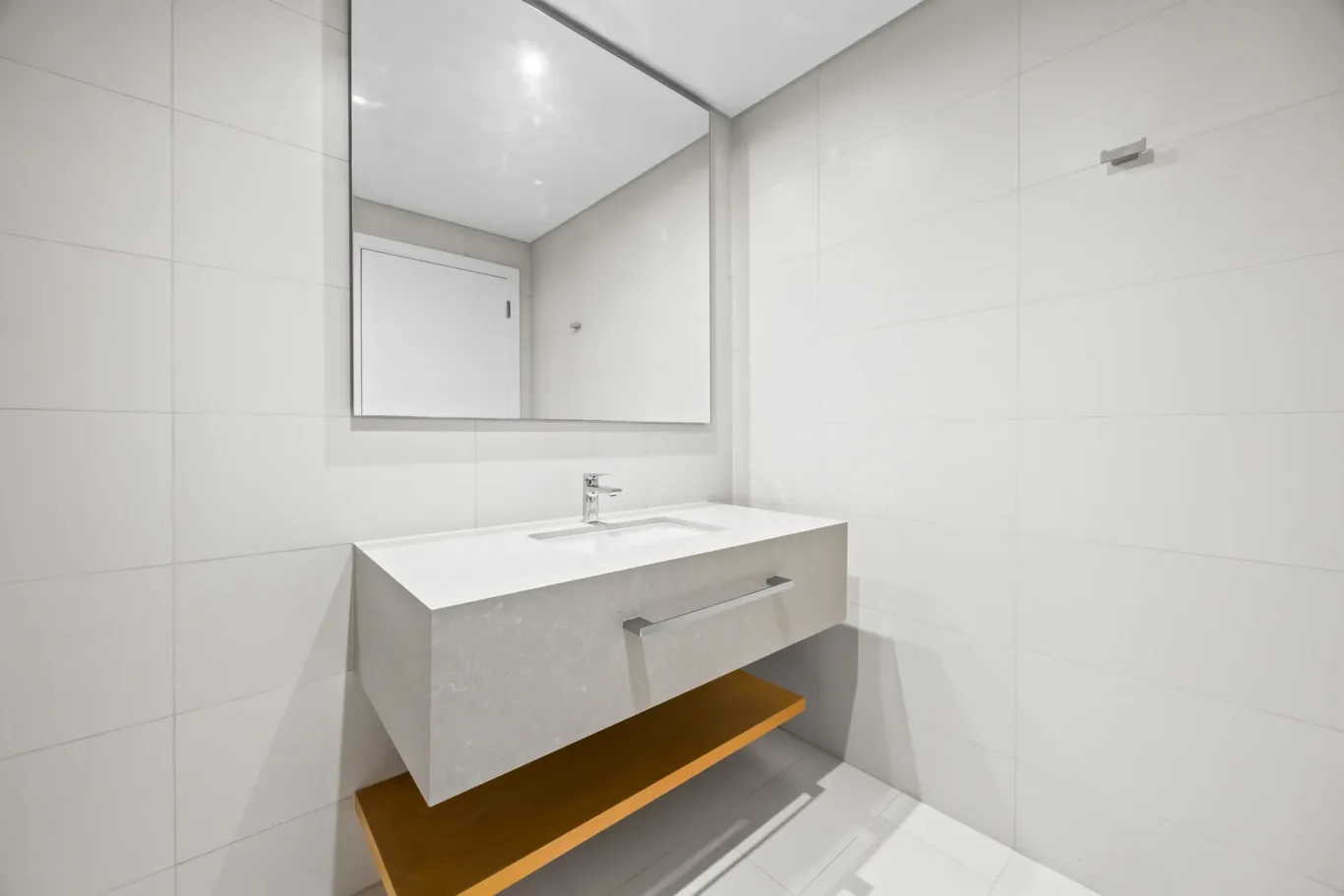 Minimalist bathroom with white tiled walls, a rectangular mirror, and a floating concrete vanity with a chrome faucet and wooden shelf underneath