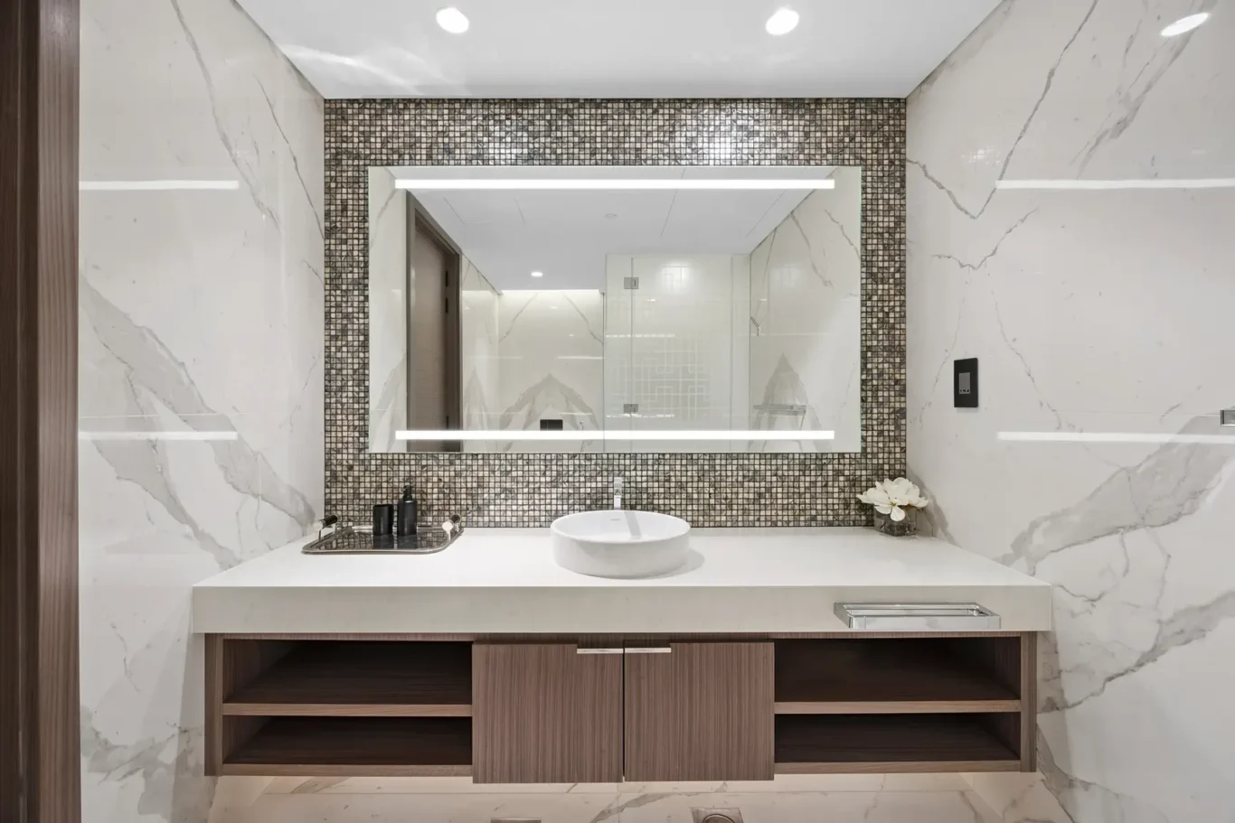 Contemporary bathroom vanity with a white vessel sink, dark wood cabinets, and a mosaic-framed backlit mirror set in marble walls.