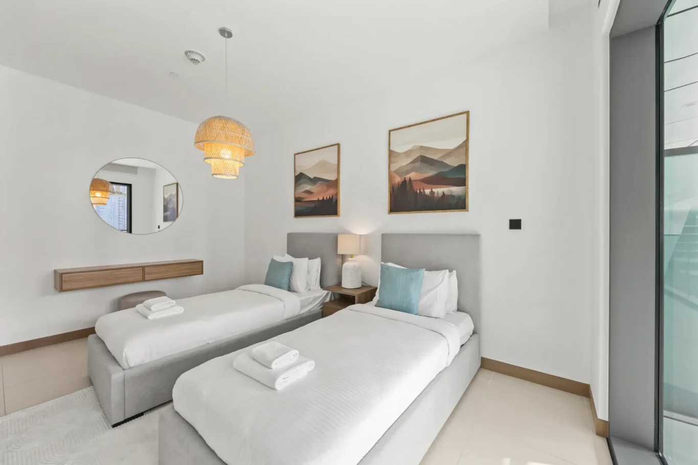 Two twin beds with white linens and teal pillows in a bright, modern hotel room with a round wall mirror and wooden console.