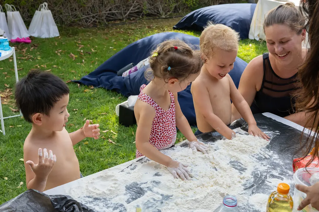 Children outdoors at a sunny backyard table, kneading flour with their hands while an adult watches and smiles nearby.