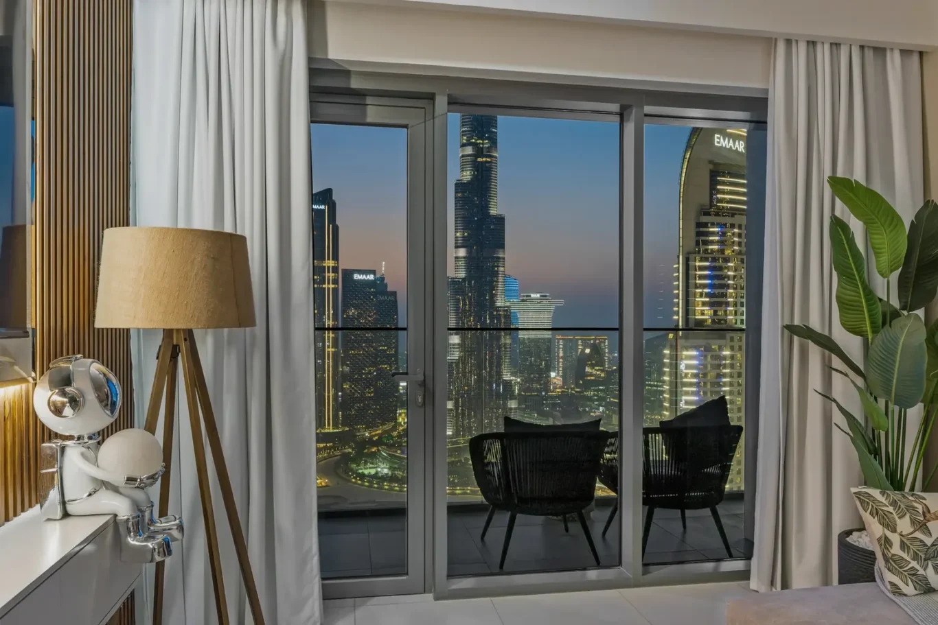 Living room with glass doors to a balcony overlooking a city skyline at dusk, indoor plant on the right and a chrome astronaut statue beside a tripod lamp on the left.