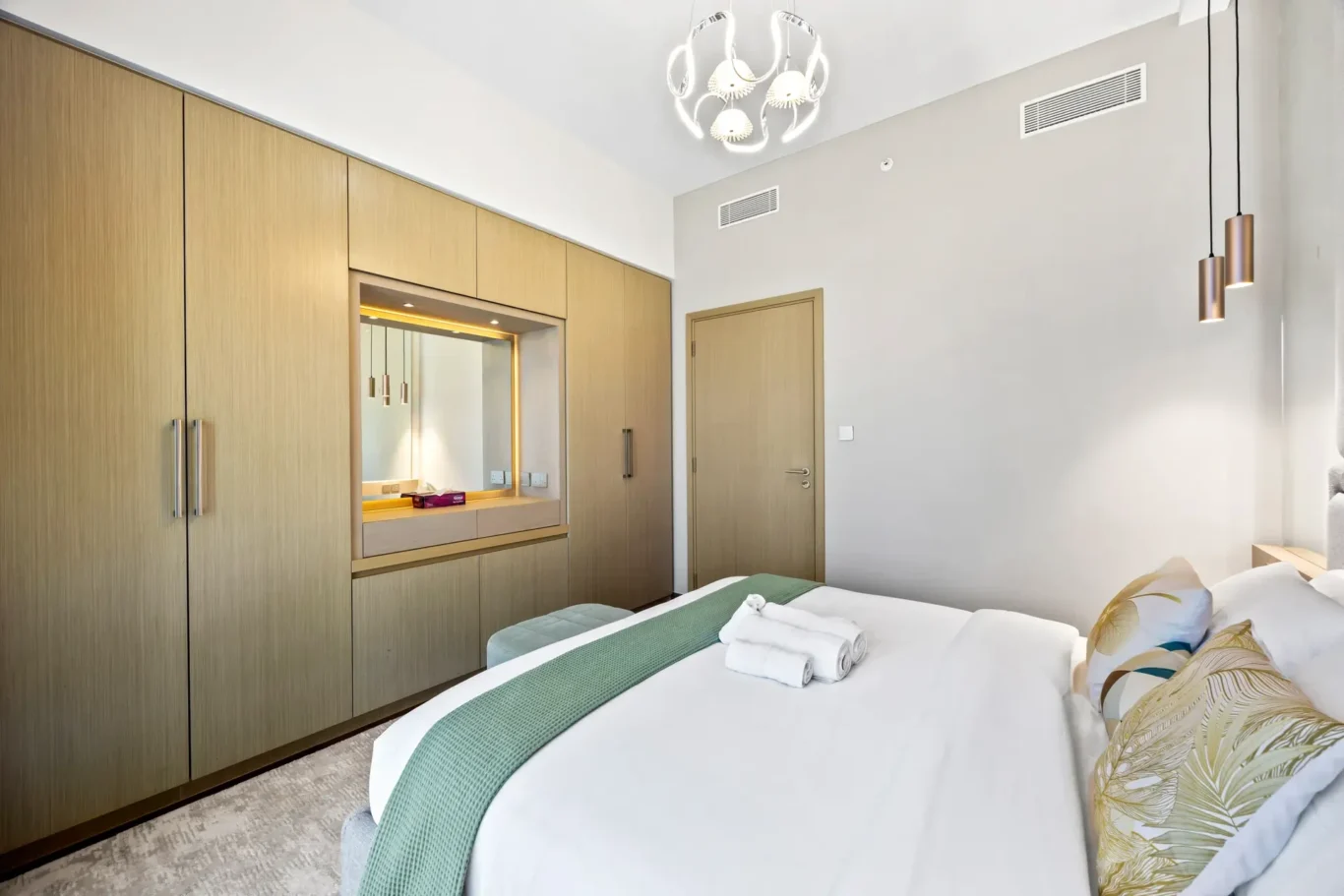 Modern hotel bedroom with wood-paneled wardrobes, a backlit vanity mirror, and a bed dressed in white with a green runner and botanical pillows.