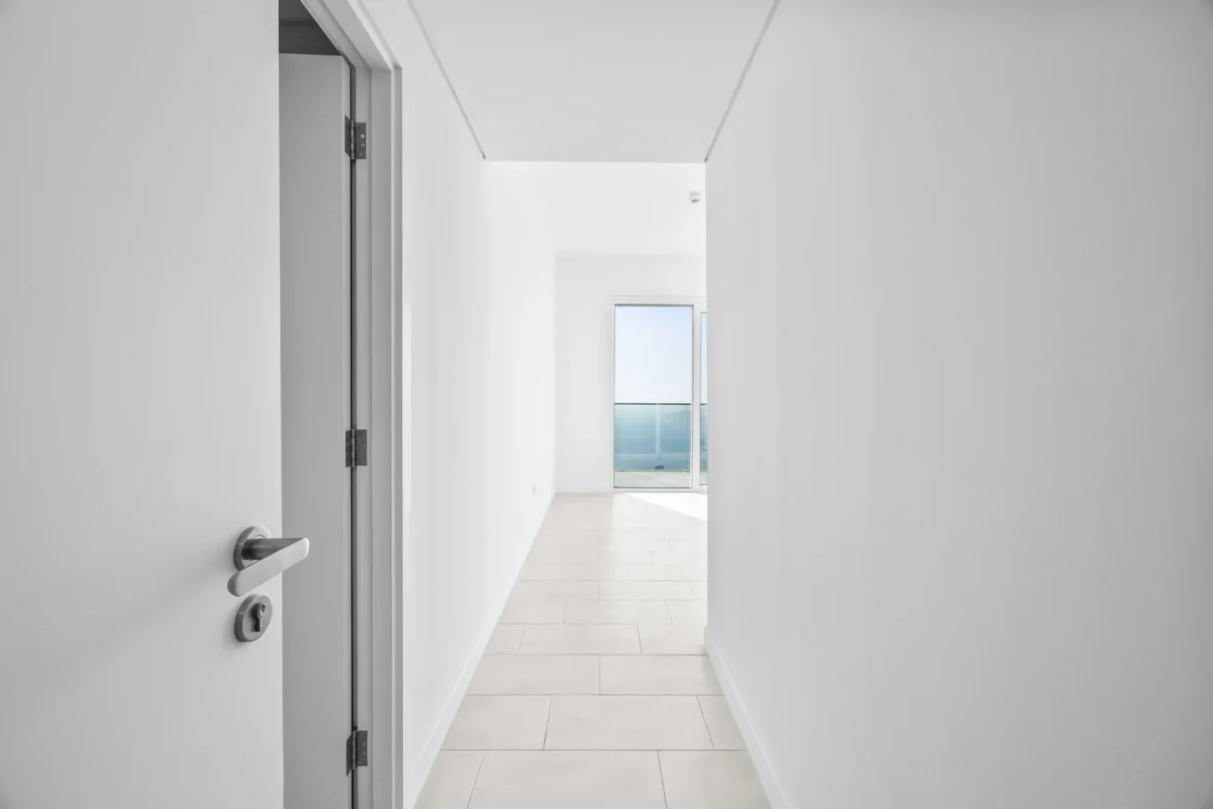 Bright white hallway with an open door on the left and a glass door at the far end showing a balcony and blue sea beyond.