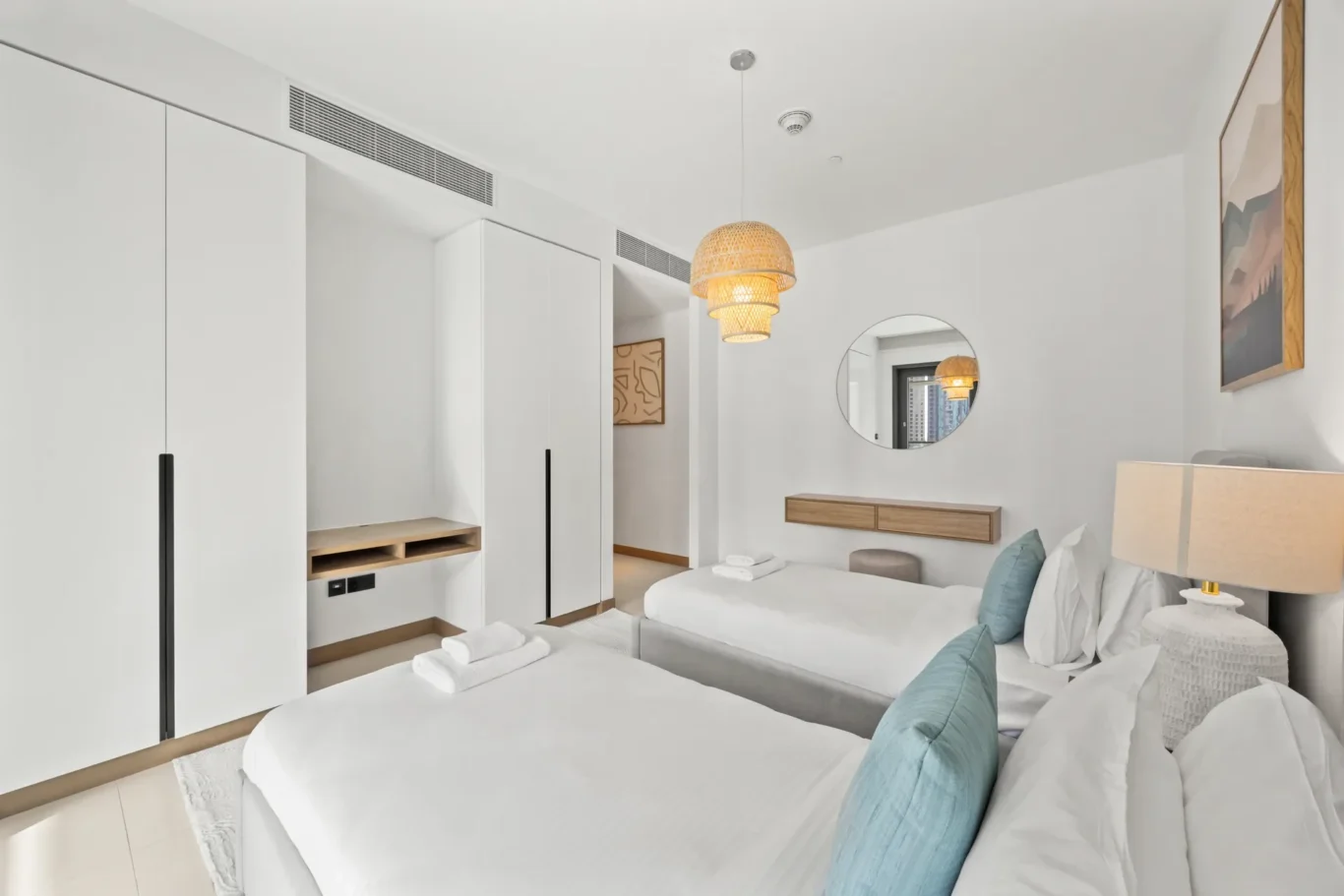 Bright minimalist hotel room with two neatly made twin beds, teal pillows, white wardrobes, and a woven pendant lamp above.