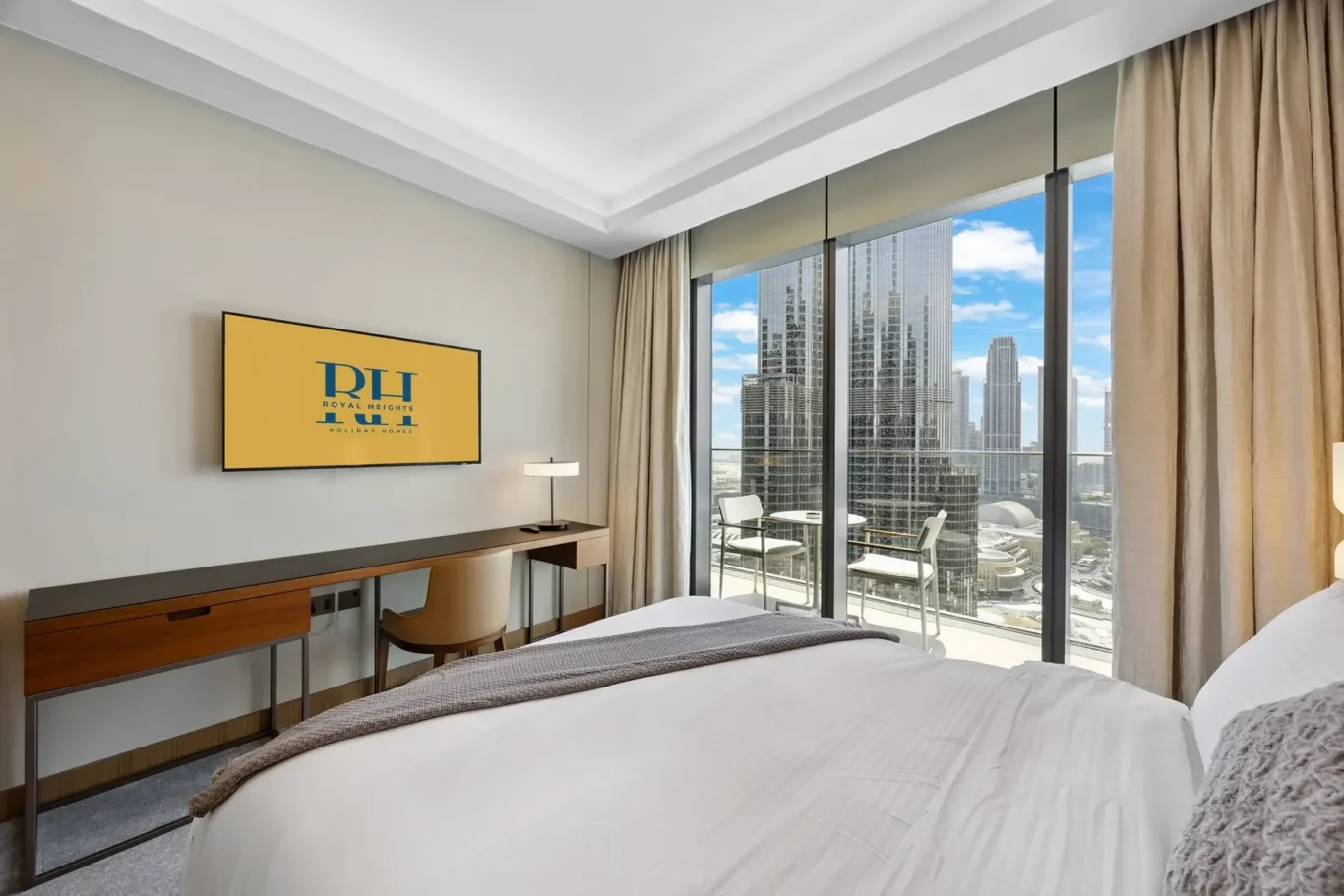 Hotel room with a large glass wall offering a city skyline view, a work desk, and a mounted yellow Royal Heights TV screen on the wall above the desk.