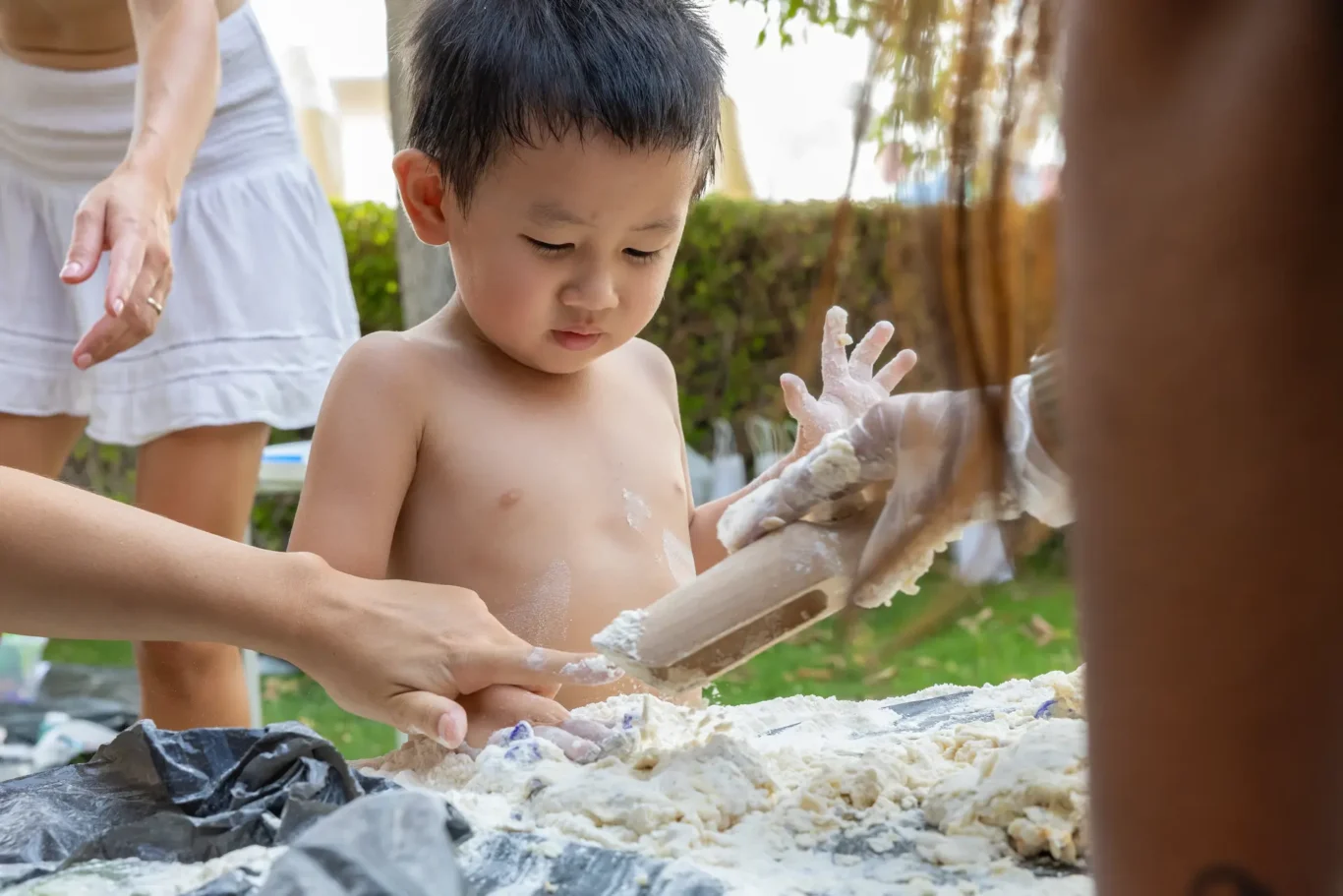 Young shirtless boy outdoors, concentrating as adults help him shape white dough or clay on a wooden surface, hands covered in paste.