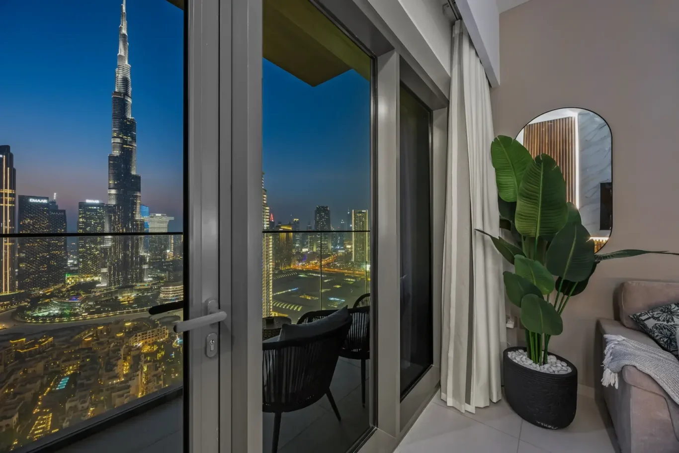 City skyline at night viewed through glass balcony doors from a modern living room, with curtains and a large potted plant nearby.