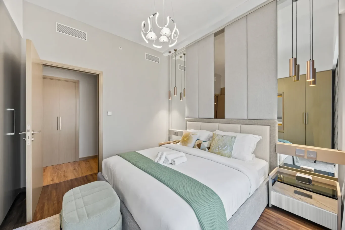 Modern hotel bedroom with a large upholstered bed, green throw, and decorative cushions; mirrored wall and hanging pendant lights create a luxe atmosphere.