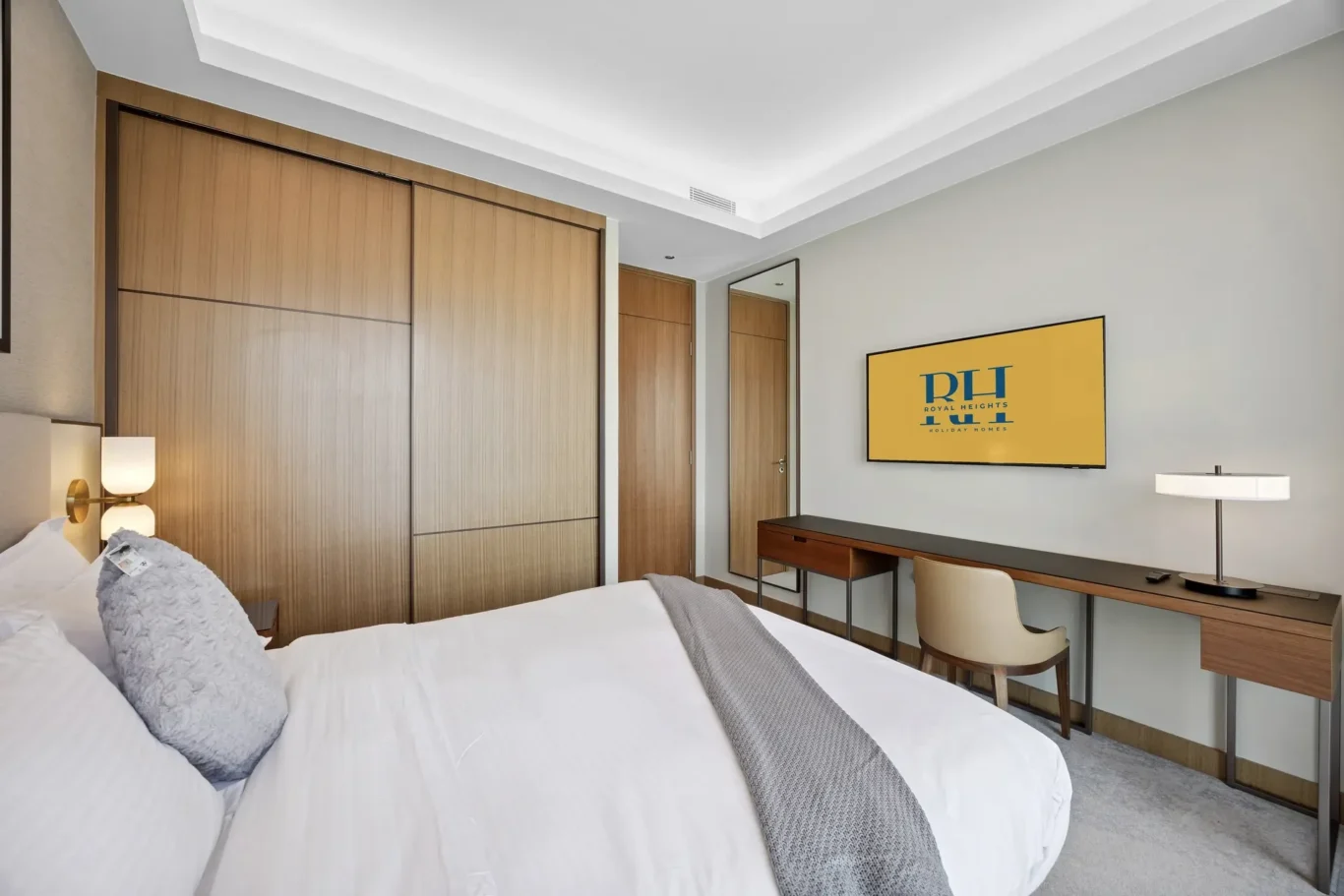 Hotel bedroom with wooden built-in wardrobes, a long desk, and a wall-mounted TV showing a yellow screen; neutral tones and modern decor.